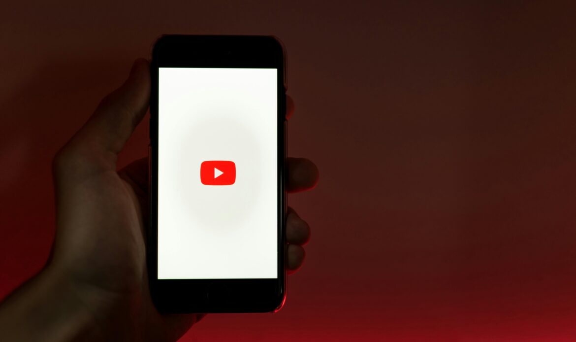 A hand holding a smartphone displaying the YouTube app against a red background.